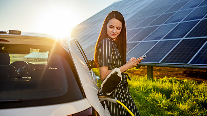 Frau, vor einem Solarpanel, an einem E-Auto lehnend